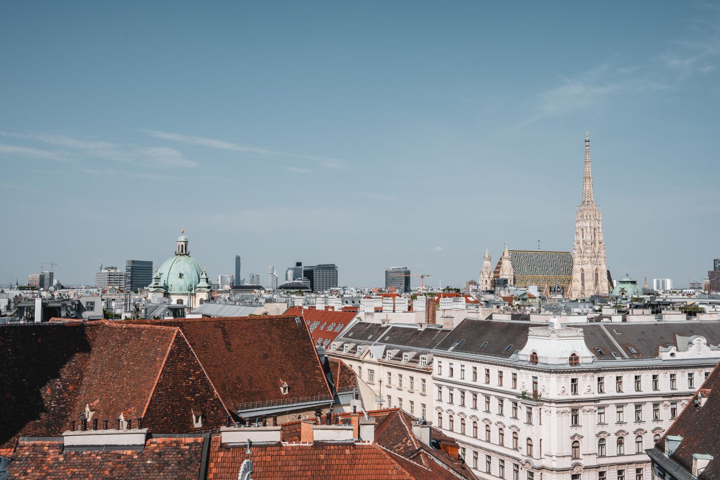 Blick über die Dächer von Wien mit dem Stephansdom und dem grünen Kuppeldach einer Kirche im Vordergrund, moderner Skyline im Hintergrund und klarem Himmel.