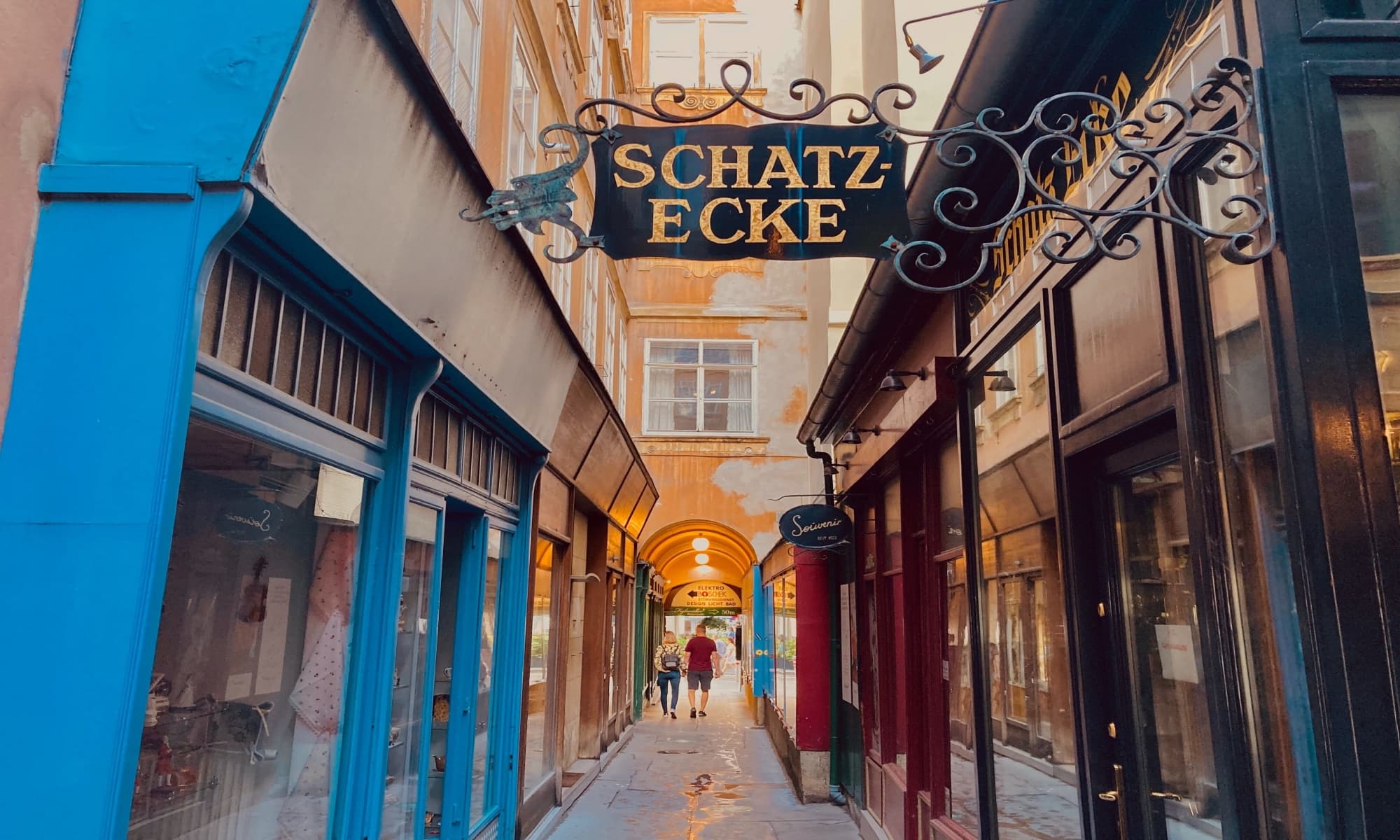 Schmale Gasse in Wien mit dem Schild "Schatz-Ecke" über dem Eingang, flankiert von bunten Ladenfassaden und einer Arkade im Hintergrund.