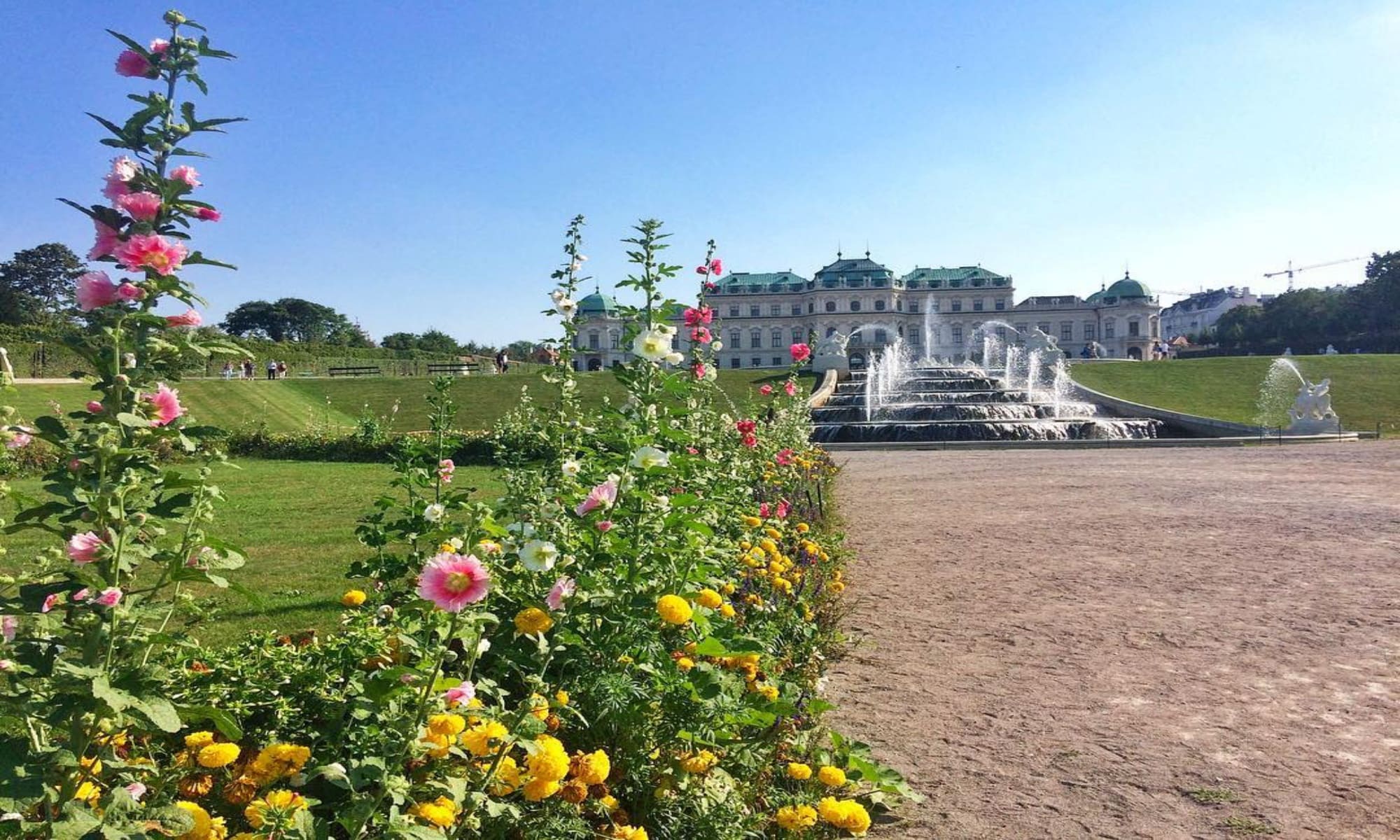 Blumenbeet mit bunten Blumen im Vordergrund, dahinter ein mehrstufiger Springbrunnen und das barocke Schloss Belvedere in Wien bei sonnigem Wetter.