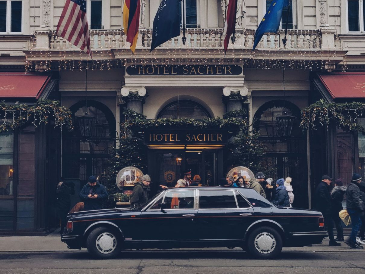 Der elegante Eingang des Hotels Sacher in Wien, geschmückt mit Lichterketten und Fahnen, sowie einem schwarzen Luxusauto im Vordergrund.