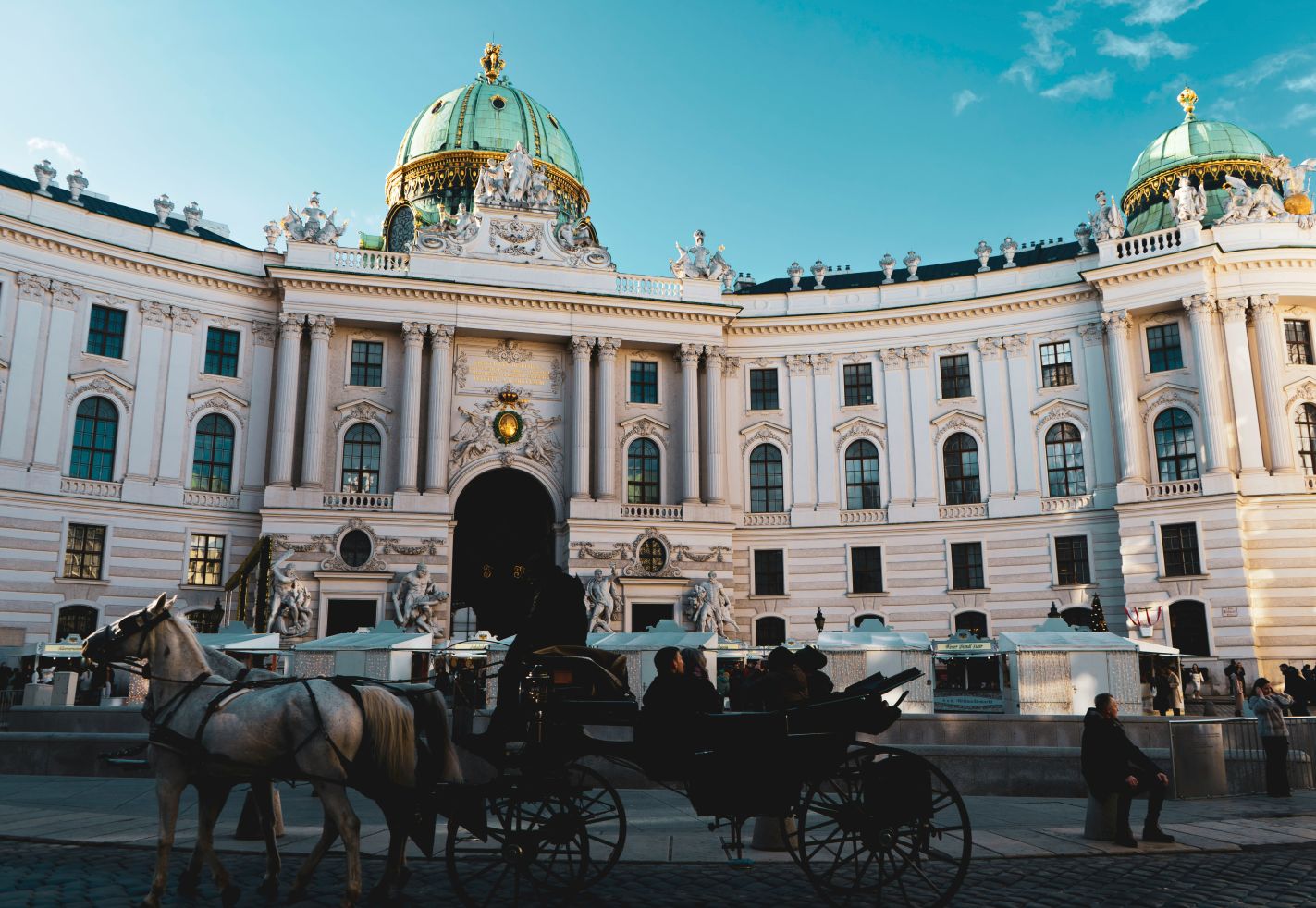 Hofburg Wien bei Tageslicht, im Vordergrund eine Pferdekutsche mit zwei weißen Pferden, barocke Architektur im Hintergrund.