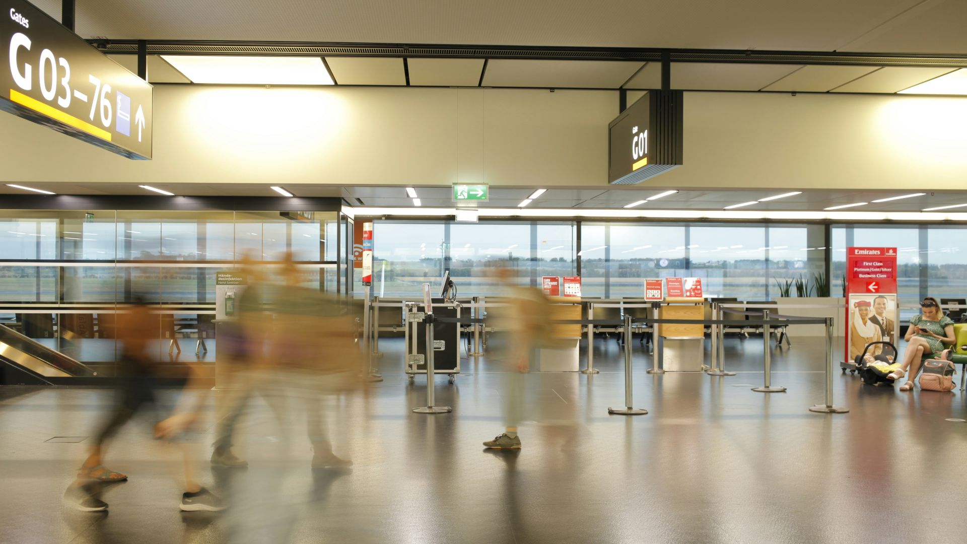 Bewegungsunscharfe Aufnahme von Reisenden im Terminalbereich des Flughafens Wien, mit Sitzmöglichkeiten und Abflugstoren im Hintergrund.