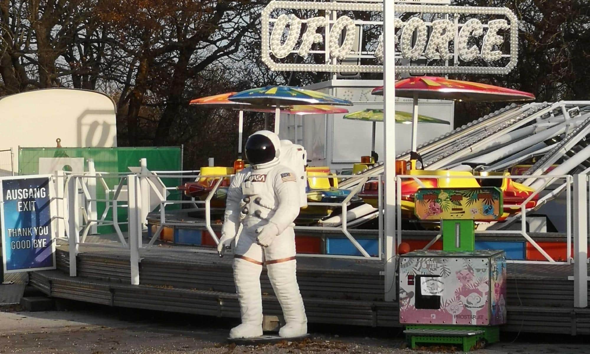 Astronautenfigur in weißem Raumanzug vor dem Fahrgeschäft UFO Force mit bunten Sitzen und einem Ausgangsschild in einem Freizeitpark in Wien