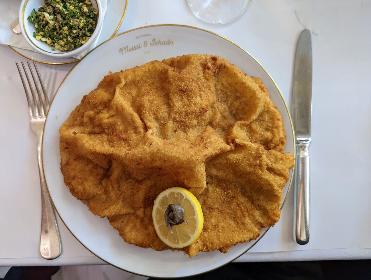 Ein klassisches Wiener Schnitzel, serviert auf einem Teller des Restaurants 'Meissl & Schadn' in Wien. Das knusprig panierte Kalbsschnitzel ist mit einer Zitronenscheibe und einer Kapernbeere garniert. Daneben liegen ein Messer und eine Gabel. Im Hintergr