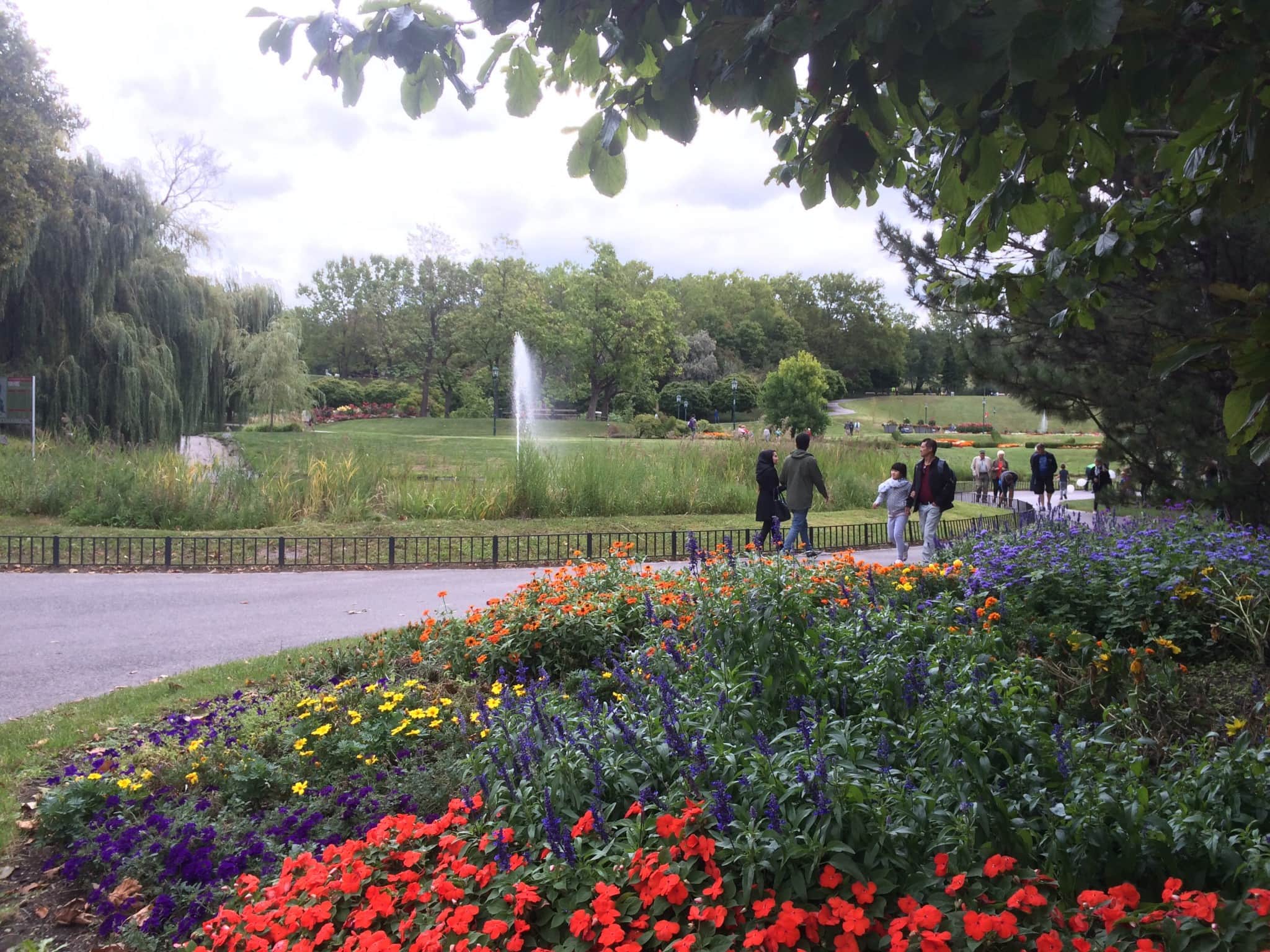 Parklandschaft in Wien mit bunten Blumenbeeten, einem Springbrunnen und Menschen, die auf den Wegen spazieren