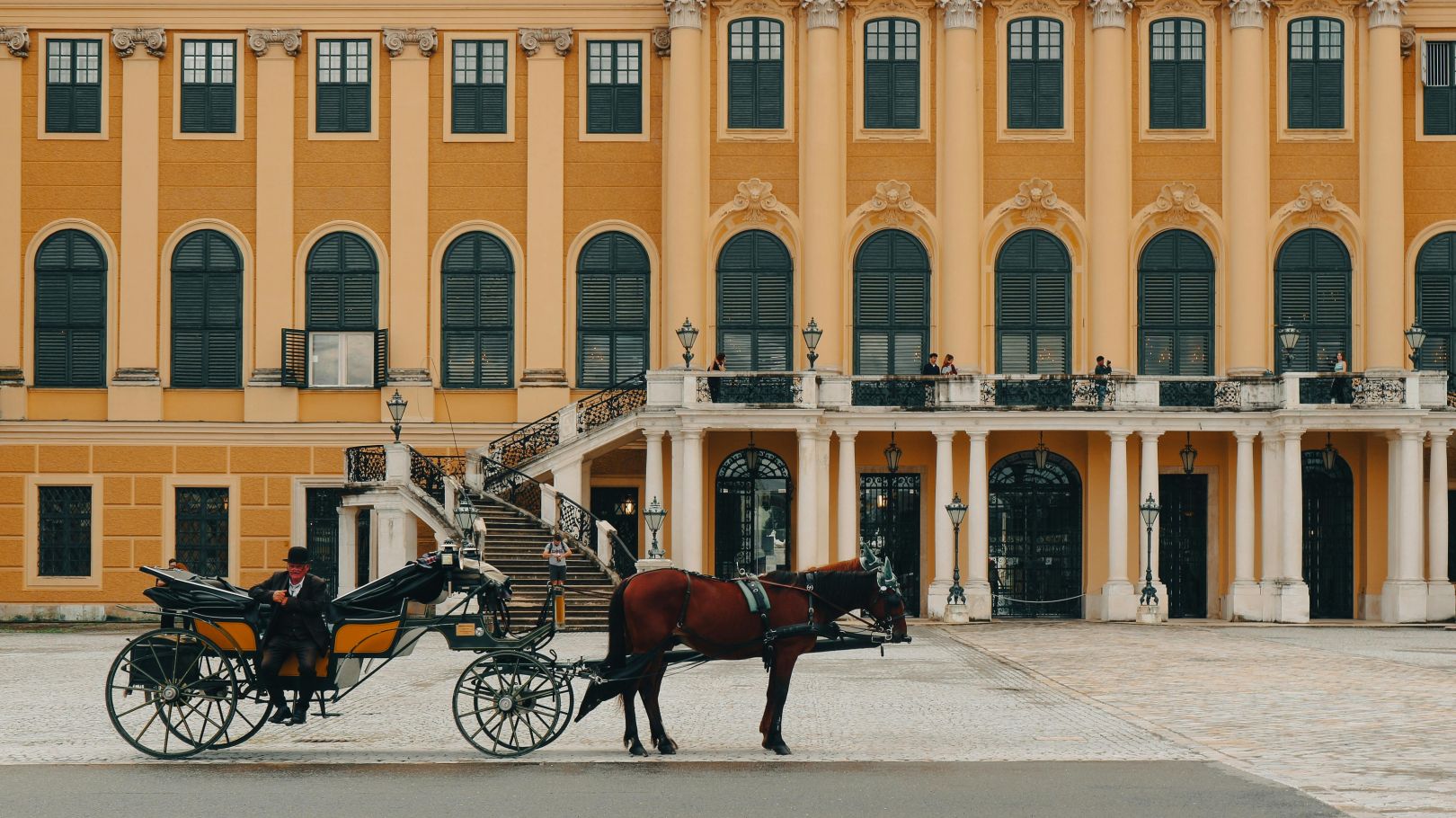 Eine traditionelle Pferdekutsche vor dem Schloss Schönbrunn in Wien, mit historischen gelben Fassaden und Besuchern auf den Balkonen.