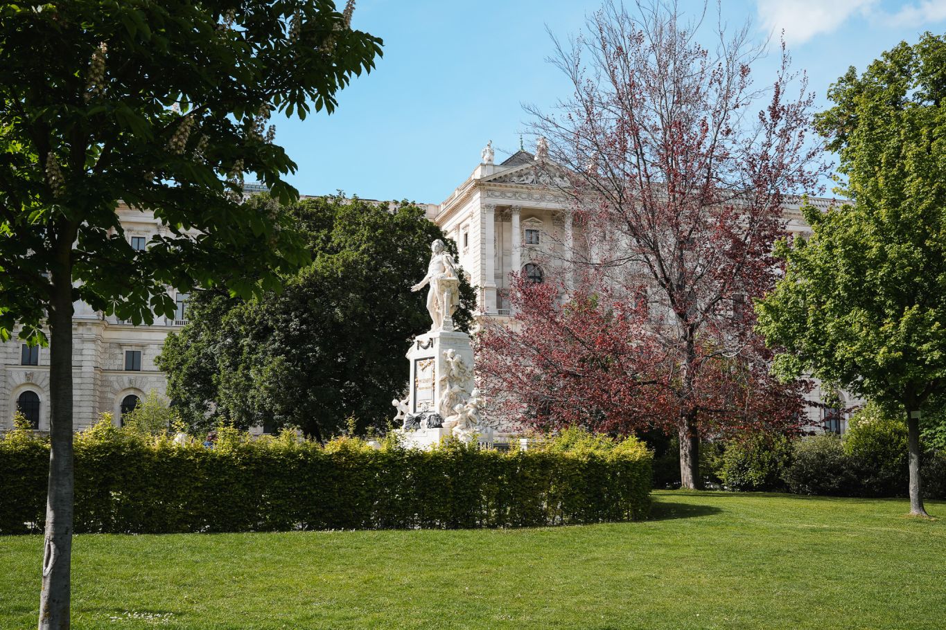 Das Mozart Denkmal im Burggarten in Wien, umgeben von blühenden Bäumen und historischer Architektur im Hintergrund.