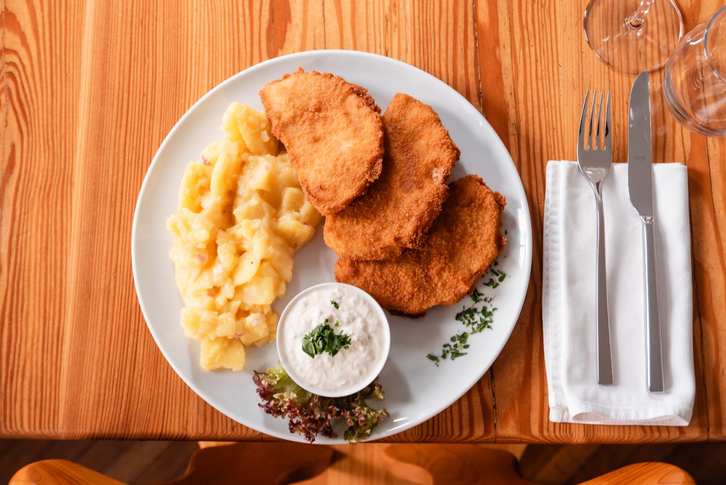 Teller mit veganem Wiener Schnitzel, Kartoffelsalat und Sahne-Dip, serviert auf einem Holztisch mit Besteck und Glas daneben. 